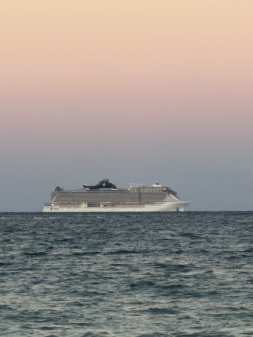 A cruise ship passing on the horizon under a soft pink sky.